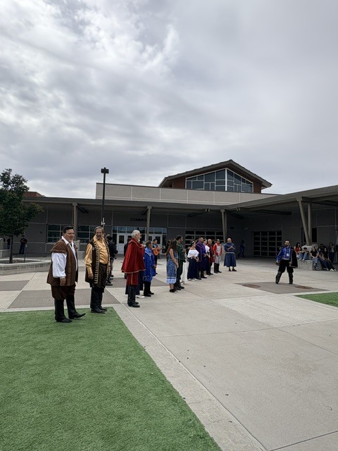 People dressed in traditional attire stand in front of a modern building with a cloudy sky.