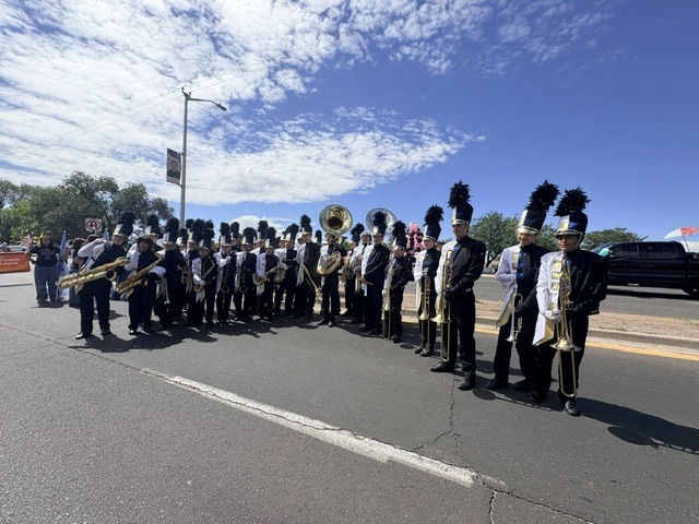 A marching band stands in a street, with musicians holding instruments and spectators nearby.
