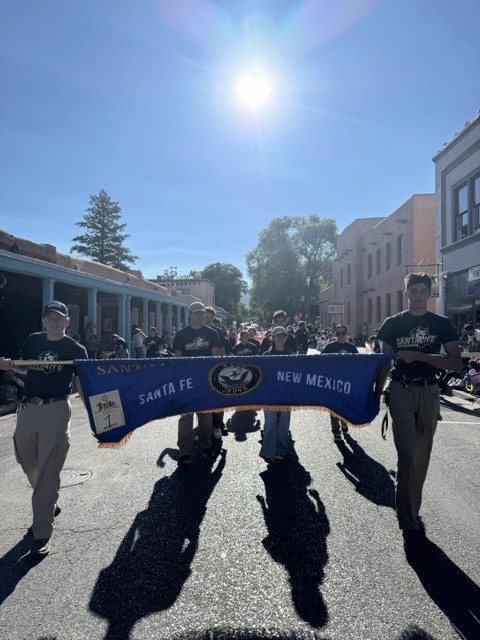 A group of people carry a banner reading "SANTA FE" and "NEW MEXICO" in a parade.