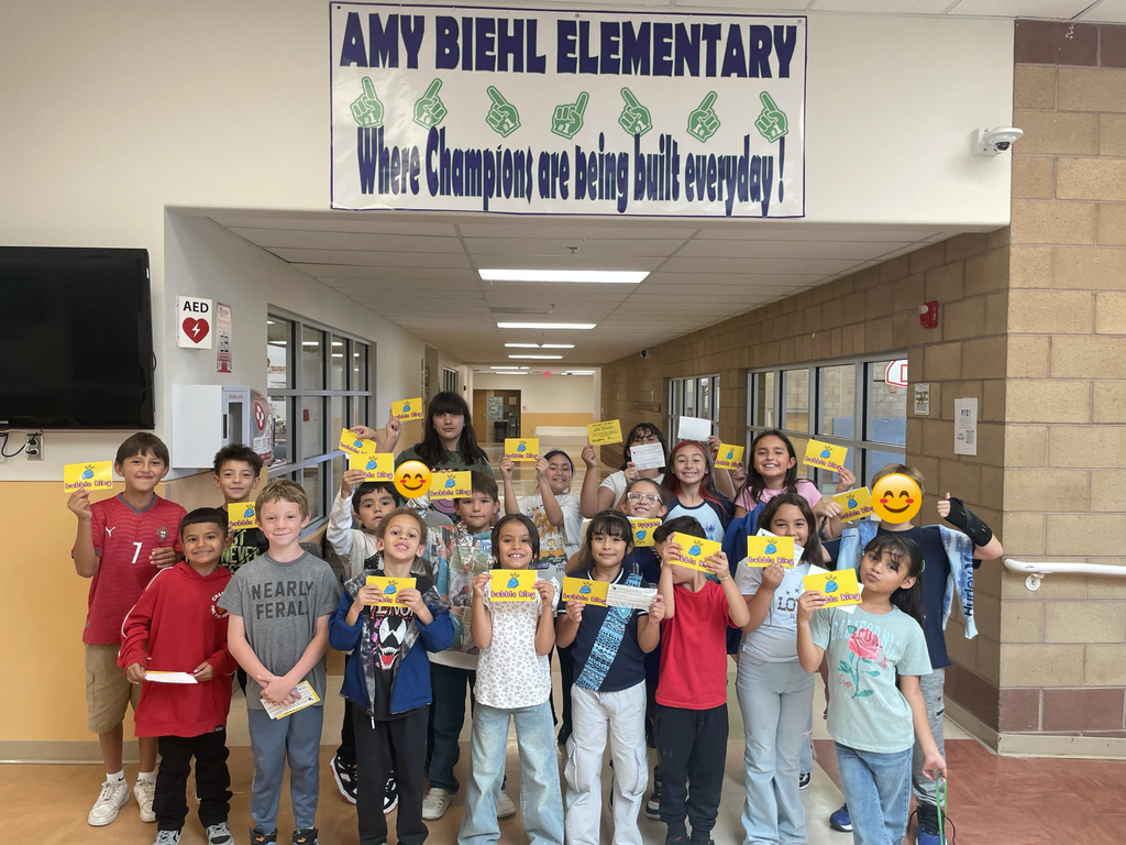 A hallway with a sign reading "Amy Biehl Elementary". A group of students holds yellow cards, smiling for a photo.