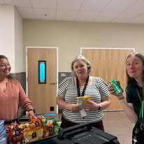 Three women near a table with snacks, one holding a can. Behind them, two closed doors and a light.