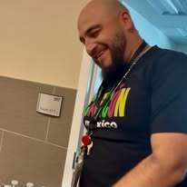 Man smiling and looking to the right, wearing a dark blue shirt with multicolored text. Tiled wall in the background.