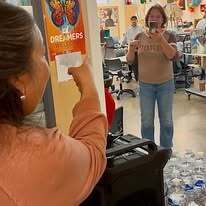 A woman points at a poster while another woman behind her takes a selfie. Bottles and chairs are visible in the background.