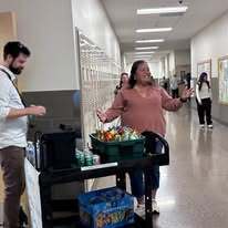 A person talks to a woman with a cart holding food in a school hallway.