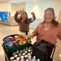 Two women are smiling, one holding a small item, standing by a table with snacks, drinks, and a speaker.