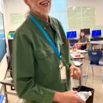 A man in a green shirt smiles, holding an item. Behind him, a classroom with desks and chairs is visible.