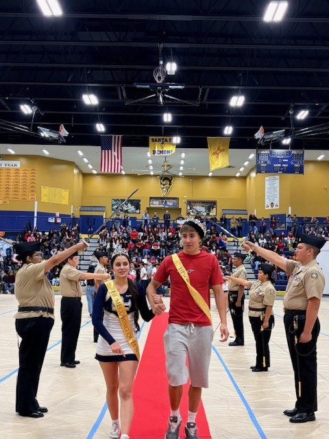 People wearing uniforms, some with hats, walk on a red carpet in a gym; some hold swords.
