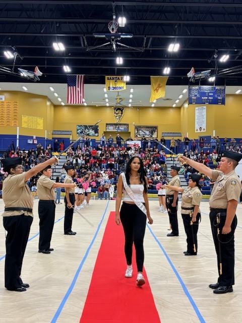 A woman walks on a red carpet in a gymnasium with uniformed adults on either side.