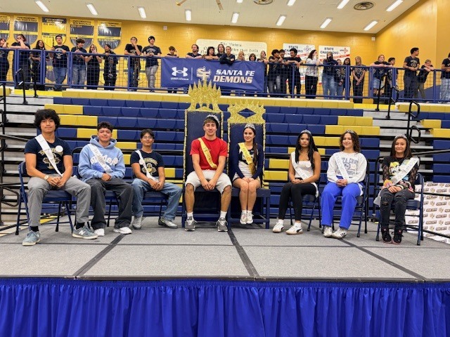 Students on a stage, seated in chairs, with several people standing in the bleachers behind them.