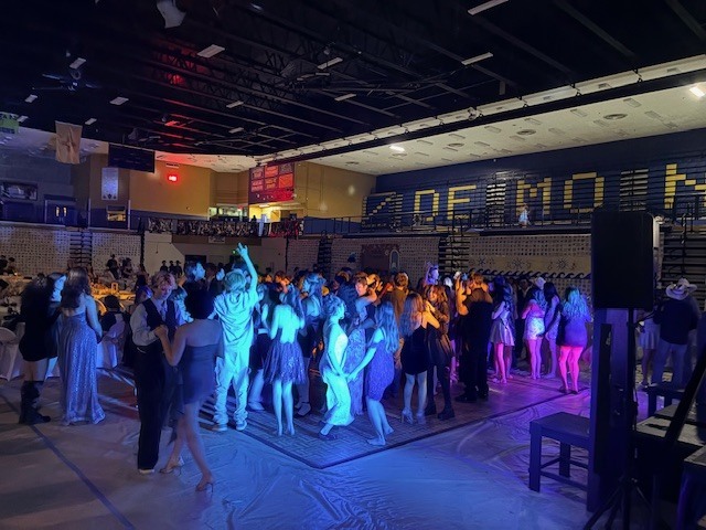 A large group of people dance in a gymnasium with blue and purple lighting.
