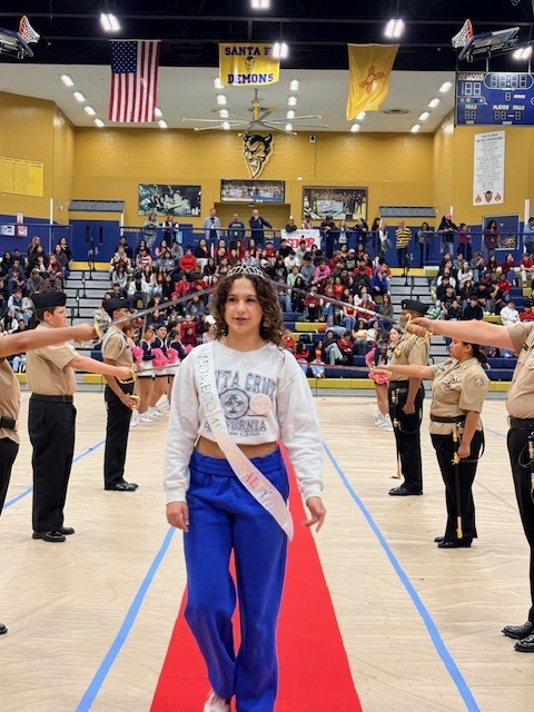 A woman walks a red carpet in a gymnasium as several people in uniform stand at the side.