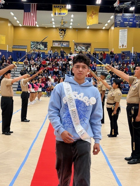 A young man walks on a red carpet, wearing a sash. Behind him are military-uniformed adults holding swords.