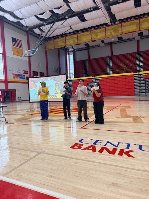 People standing in an indoor gym, four holding cameras, one wearing a hat, floor has red and blue markings, a large screen in the background.