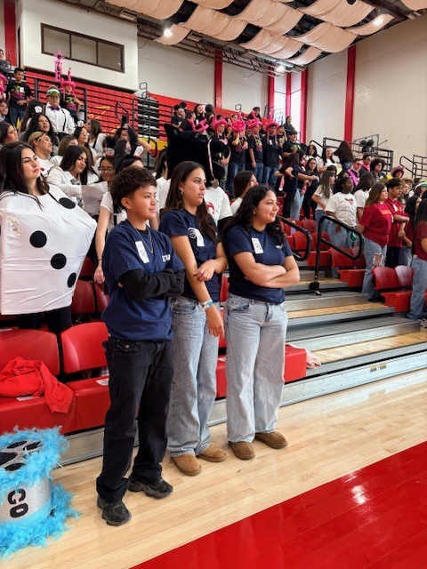 Four students in blue shirts and jeans stand on a gym floor, watching a crowd on bleachers.