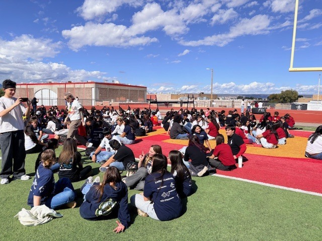 Many students sit in rows on a field; a man stands at the front and addresses the crowd.