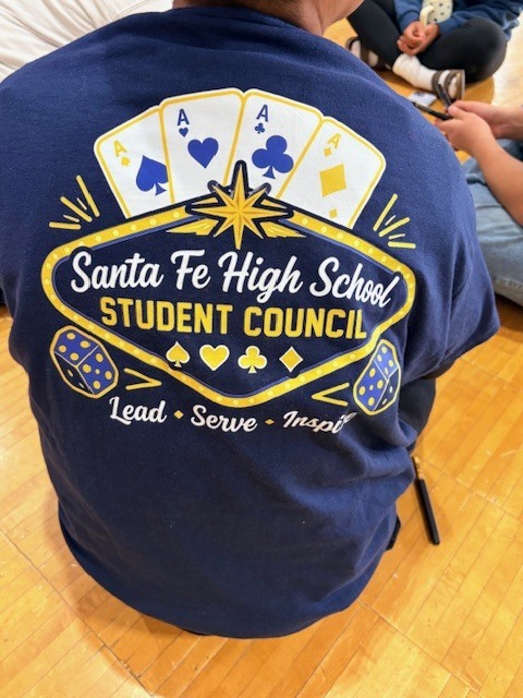 Back of navy blue T-shirt with gold design featuring cards, dice, and words. Person sits on floor.