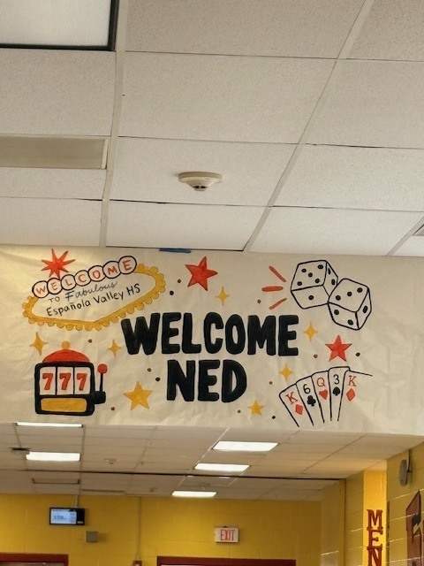 Yellow hallway with a banner reading "Welcome Ned" with dice and playing cards. Bright lighting.
