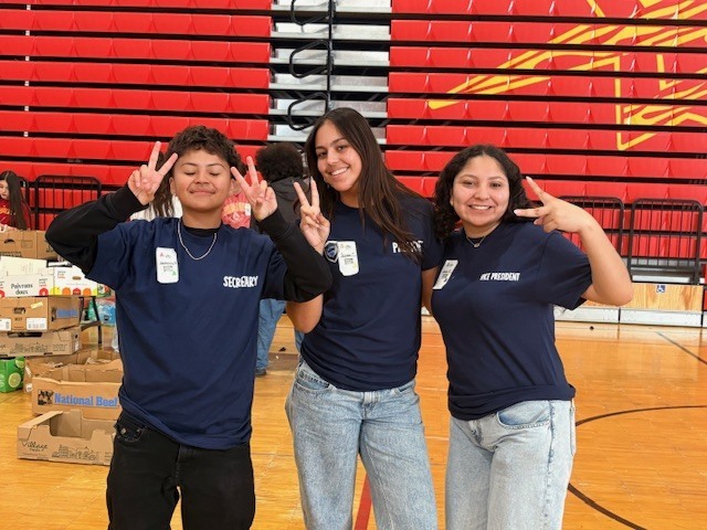 Three students pose with peace signs in a gym with red bleachers and yellow lines.