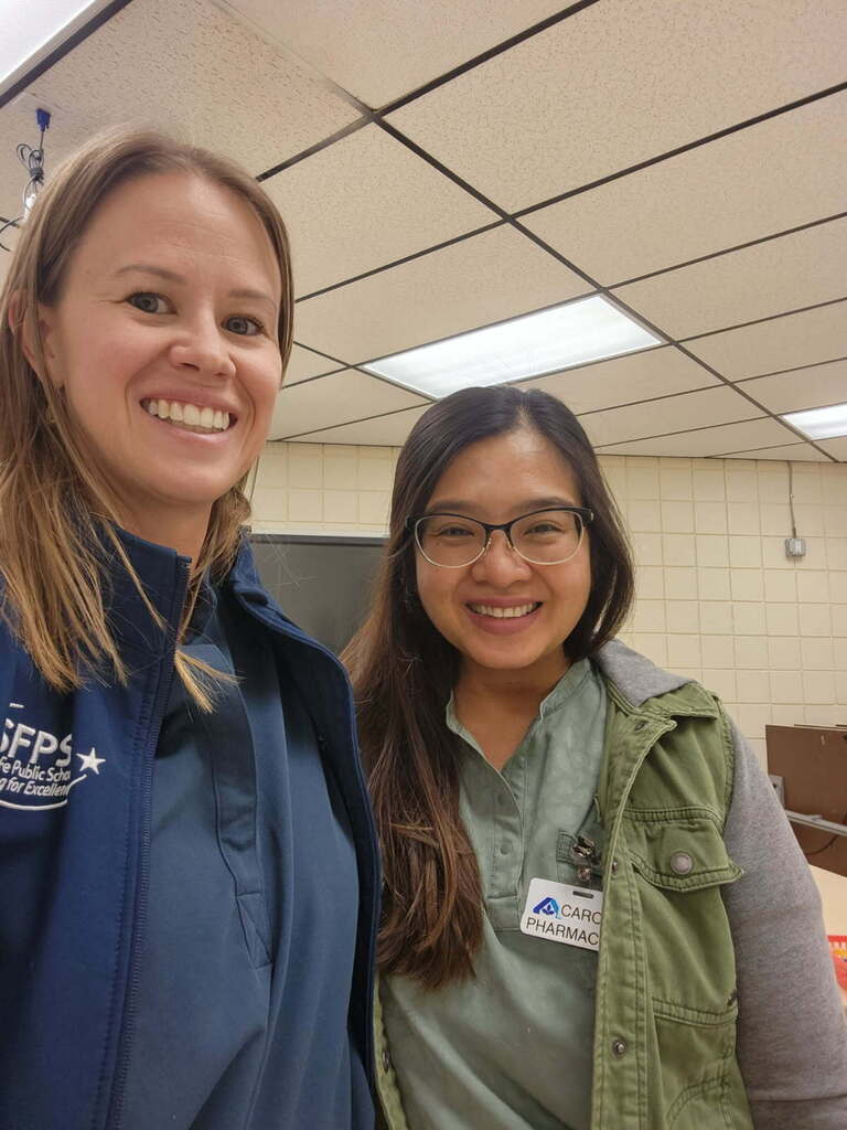 Two women smiling, one in a blue jacket, the other in a green vest, standing near a tiled wall.