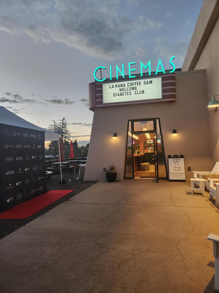 A cinema building with the sign "Cinemas" and a marquee. Chairs and a table with an umbrella.