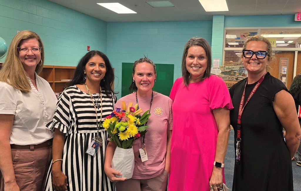 Ms. Rice, Ms. Drost, Ms. Feilner, Ms. Dorsey, and Ms. Neu lined up smiling, while Ms. Feilner receives flowers.