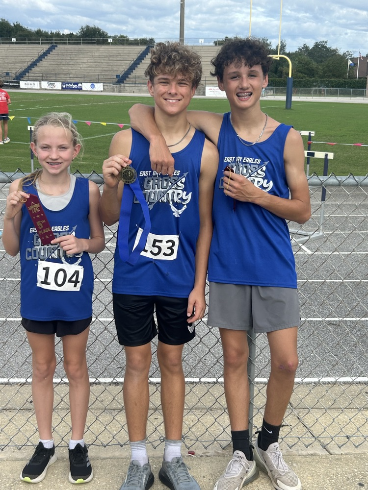 East Bay cross-country runners and coaches pose in blue team uniforms after their final meet, with several athletes holding medals and ribbons near the track.