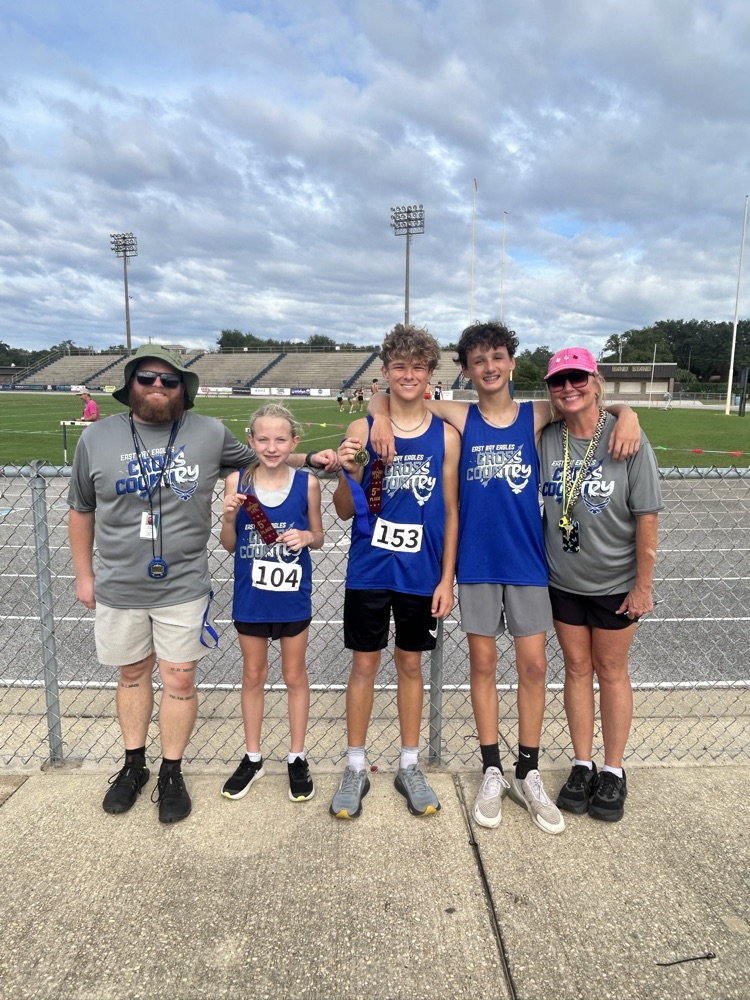 East Bay cross-country runners and coaches pose in blue team uniforms after their final meet, with several athletes holding medals and ribbons near the track.