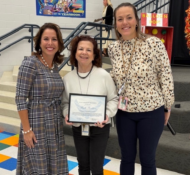 1: Three women pose in the school atrium, with the woman on the left holding a framed staff recognition certificate.