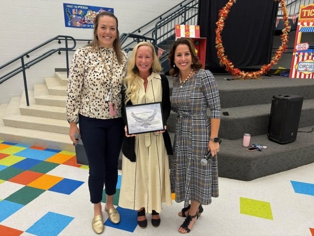 1: Three women pose in the school atrium, with the woman on the left holding a framed staff recognition certificate.