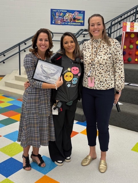 1: Three women pose in the school atrium, with the woman on the left holding a framed staff recognition certificate.