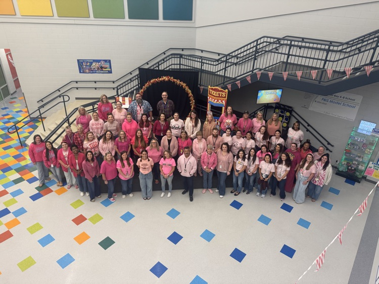 East Bay K-8 staff members dressed in shades of pink pose together in the school atrium beneath the staircase.