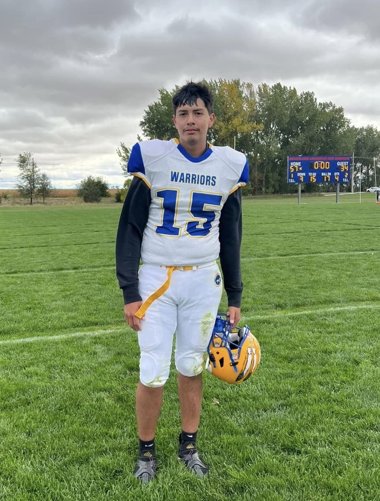 Number 15, Isaiah Yellow Cloud, stands on a grassy field, holding a yellow helmet. under a cloudy sky.