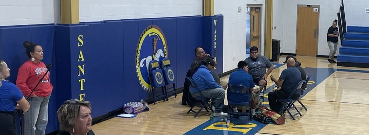 picture of the drum group sitting on chairs around their drum in the gymnasium
