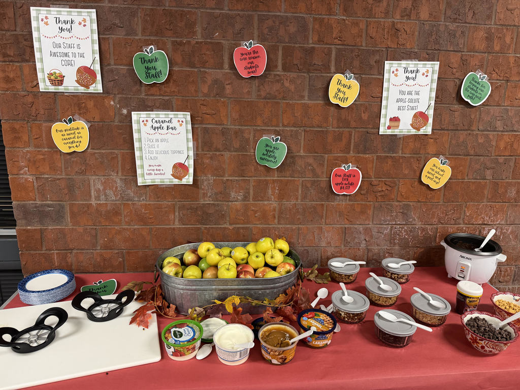 A festive “Apple Bar” setup against a brick wall decorated with colorful apple-shaped signs and thank-you posters for staff. On the table are apples in a metal tub, apple slicers, caramel dip, yogurt, and several bowls of toppings like nuts and chocolate chips. The table is covered with a red cloth and autumn leaves for decoration, creating a warm, fall-themed display.