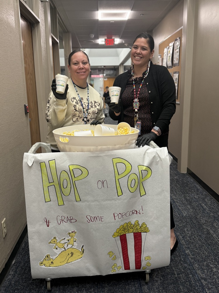 photo of two female principals holding cups of popcorn by a white sign with a popcorn bucket that reads “Hop on Pop & grab some popcorn"