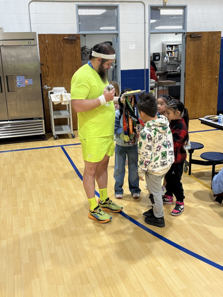 small students standing around a male teacher while he shows them his marathon medals. teacher is wearing fluorescent green workout clothing