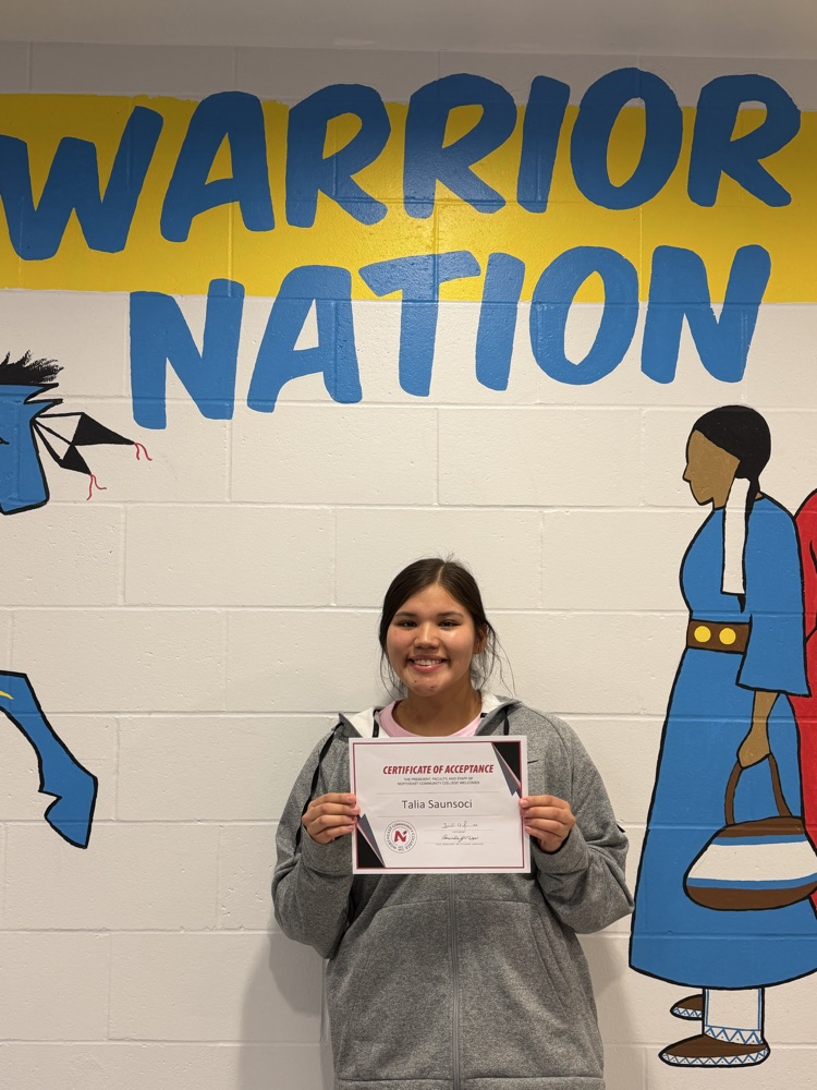 Female student in gray sweater holding up her acceptance letter. White wall behind her with a painting on it of a lady in a blue dress