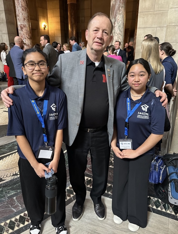 Female students dressed in all black with a male Nebraska senator dressed in black pants, black shirt with a gray blazer jacket. posing for a group picture 