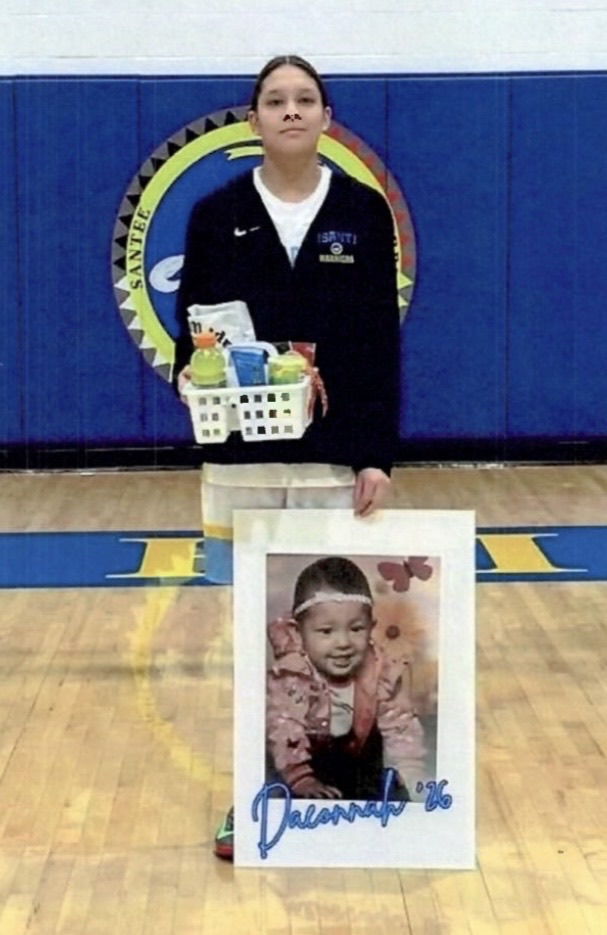 photo of a female basketball player in a white uniform with a black jacket over it. player is holding her baby picture for senior night 