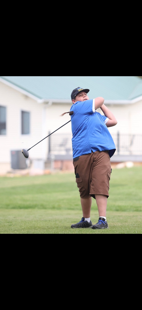Photo of a high school golf athlete swinging the club standing on a green golf course. Student has a blue shirt on and brown shorts.