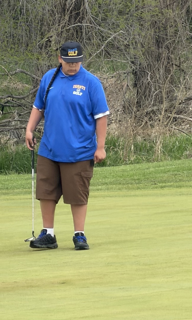 Photo of a high school golf athlete holding the club standing on a green golf course. Student has a blue shirt on and brown shorts