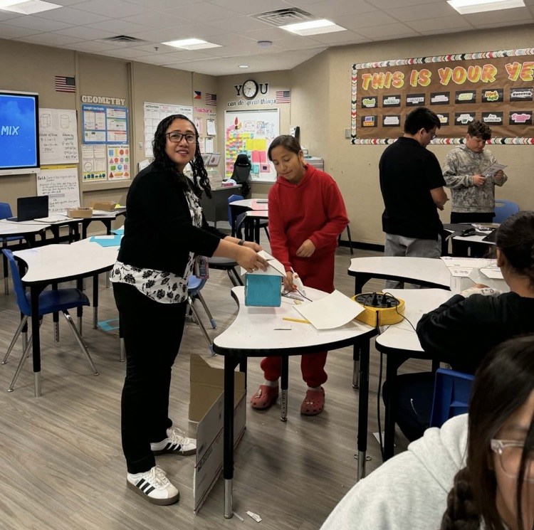 Photo of the math teacher, female, black blouse, black pants, white shoes standing by a white desk and a male student with red pants and a red sweater on