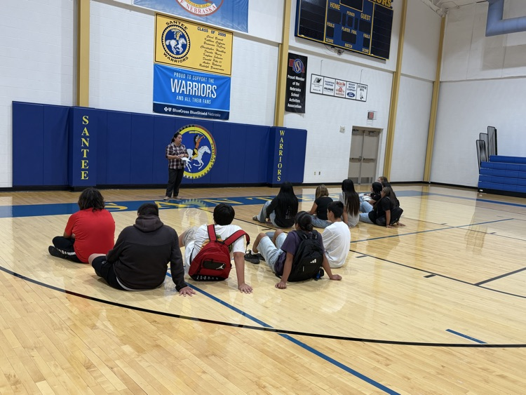 photo of students sitting on the gym floor facing the teacher listening to instructions 