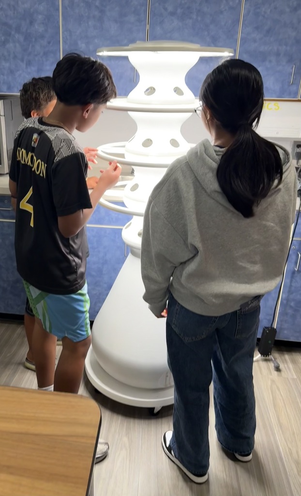 Three students work together around a brightly lit indoor hydroponic growing tower in a classroom.