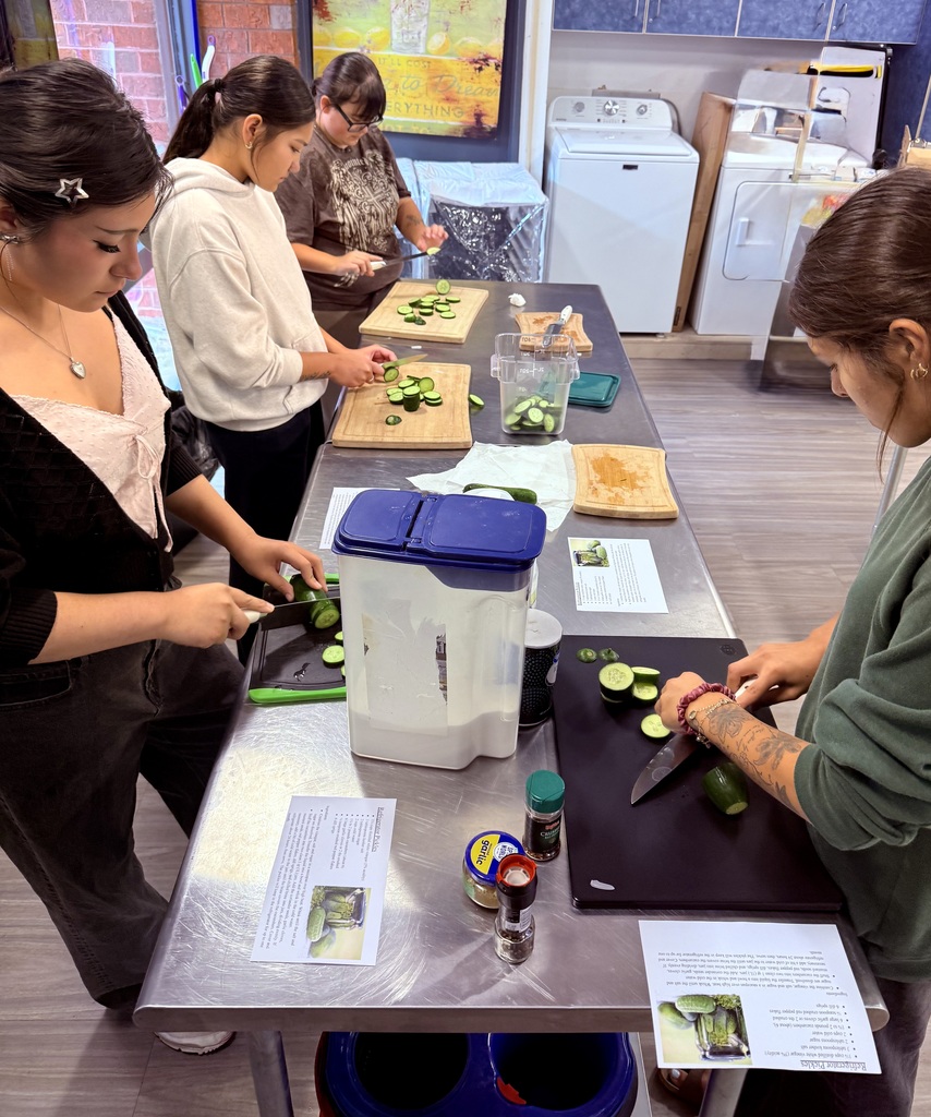 Four students slice cucumbers at a stainless steel worktable while following recipes to prepare refrigerator pickles during a hands on culinary lesson.