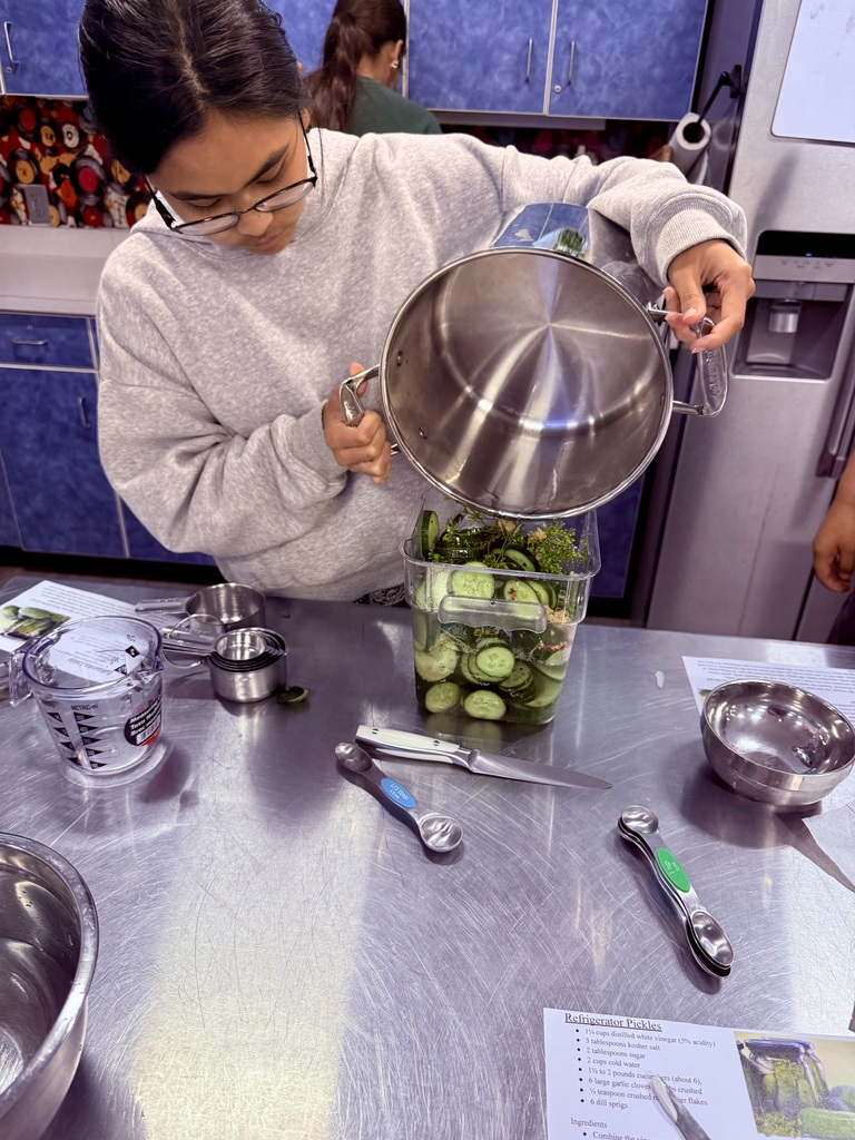 A student pours brine from a large stainless steel pot into a container filled with sliced cucumbers, garlic, and fresh dill while making refrigerator pickles in a culinary classroom.