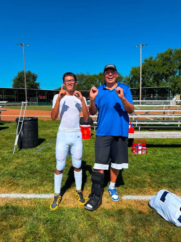 photo of a football player in all white uniform with his coach in a blue shirt holding up their fingers “1,000” for 1k passing yard record 