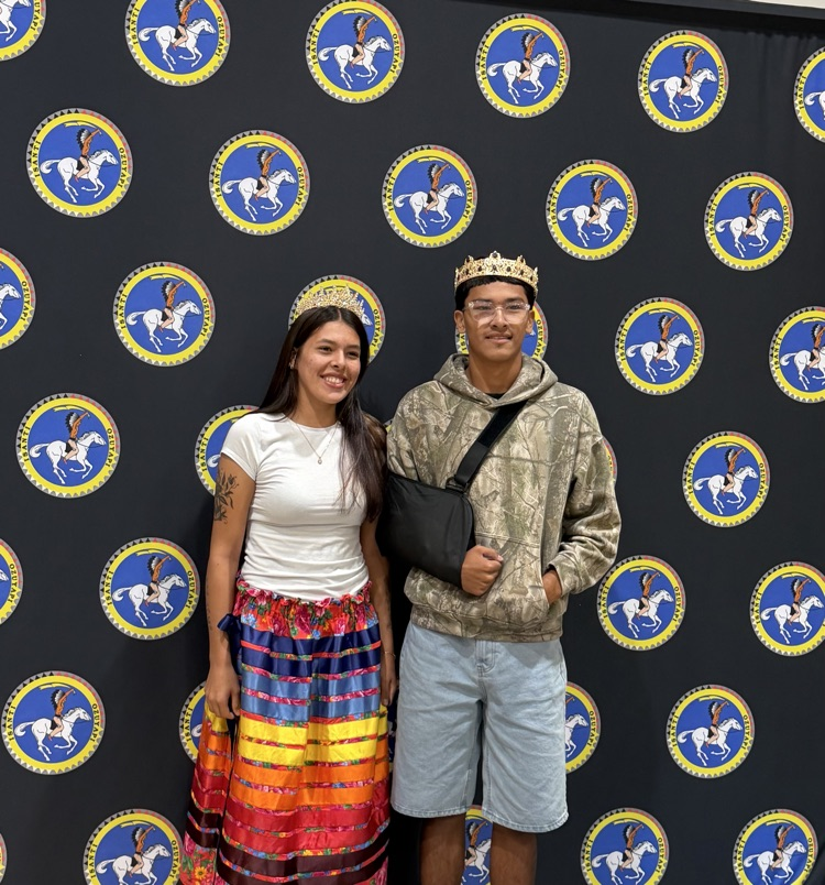 Isanti school logo background with homecoming queen in rainbow skirt and white shirt, king in a camouflage hoodie and jean shorts. both with gold crowns on