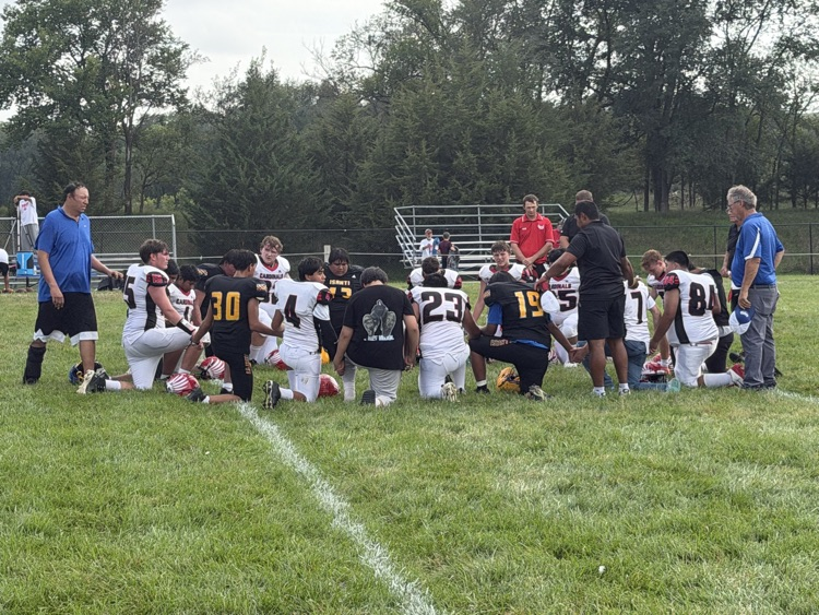 photo of opposing teams on the field. white jerseys and black jerseys. students all kneeling praying together 