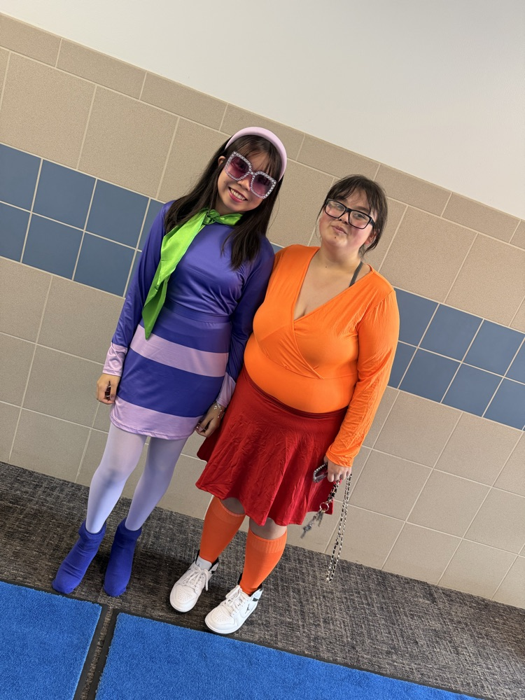 two female students in 70s costumes posing for a photo by a gray wall
